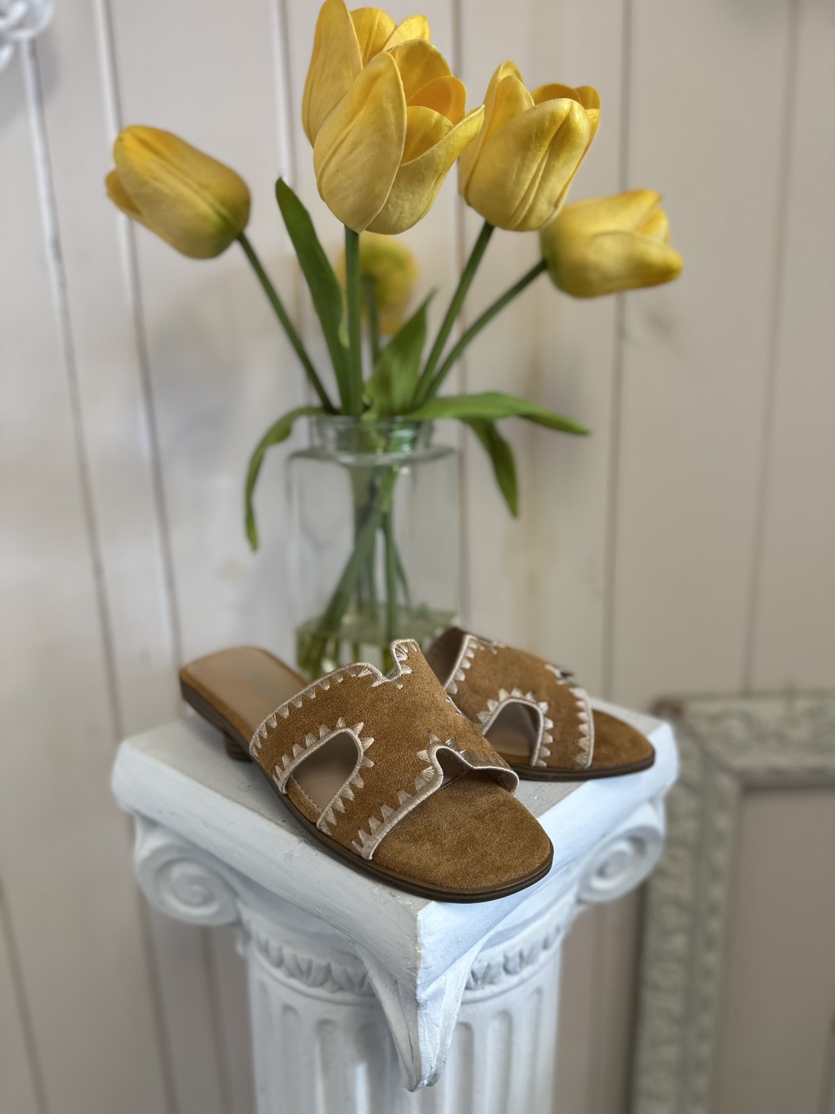 Melly Tan Shoe displayed on a pedestal with yellow tulips, showcasing trendy coastal style from Raggdoll Boutique.