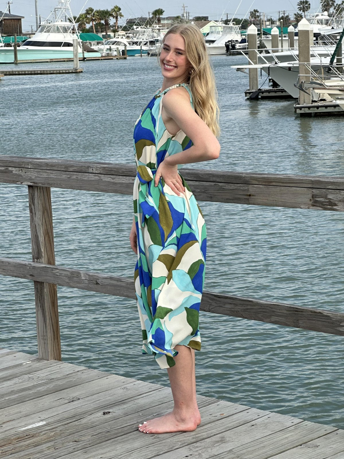 Young woman in a vibrant tropical leaves midi dress at a marina, perfect for beach parties and summer getaways.