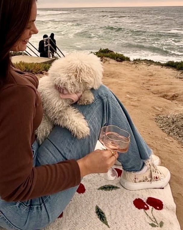 Woman enjoying wine on beach with dog in trendy Sadie Sun Sneaker by Raggdoll Boutique, perfect for coastal fashion.