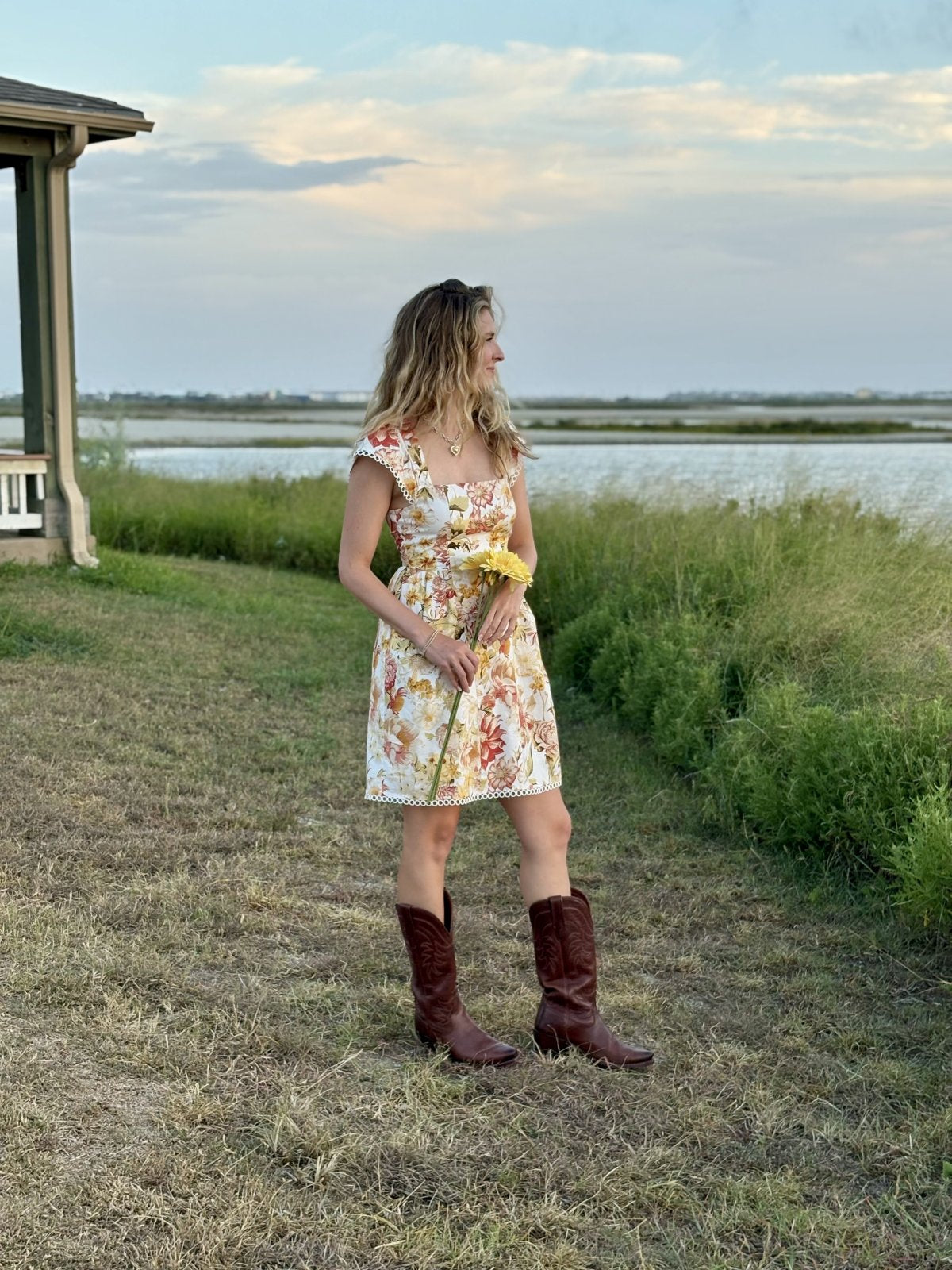 Woman in floral mini dress and brown boots standing by water, showcasing coastal boutique clothing style and boho-chic vibes.