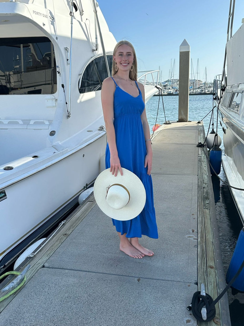Woman wearing a Brielle Dress in cobalt blue on a dock by a marina, holding a wide-brimmed hat.