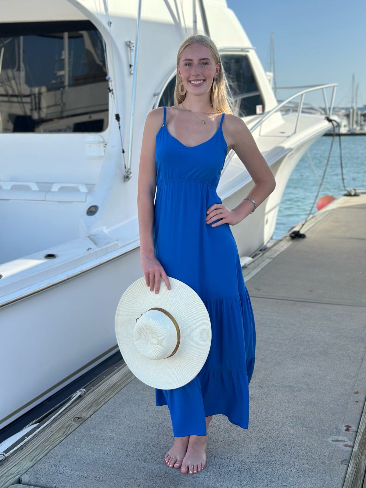 Model wearing the Brielle Dress, an elegant cobalt blue maxi dress, by the marina with a sun hat.