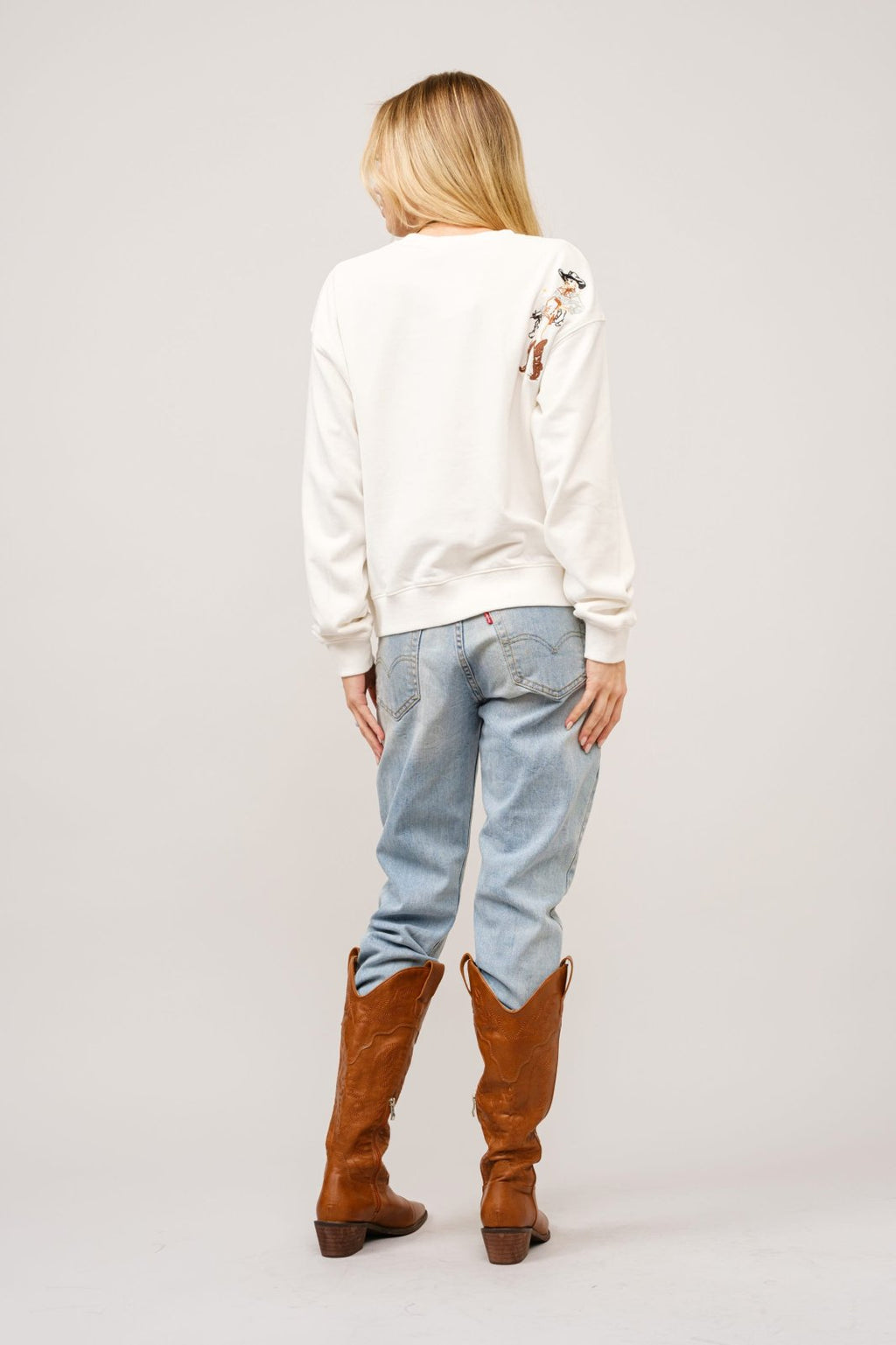 Back view of a woman wearing a cute western motif embroidered sweatshirt, paired with jeans and brown boots. Perfect for coastal boutique fashion.