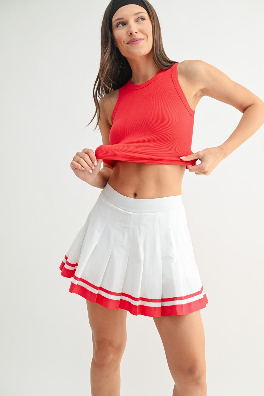 Model wearing a ruby muscle tank with a white pleated skirt, showcasing coastal boutique clothing and stylish tops for women.