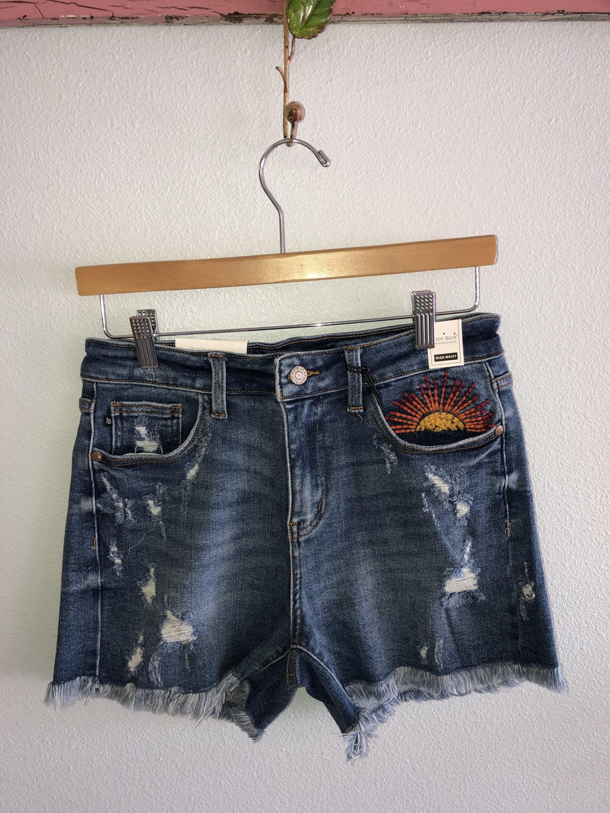 JUDY BLUE HI-RISE SUN EMBROIDERY CUTOFF SHORTS hanging on a rack, showcasing trendy coastal style from Raggdoll Boutique.