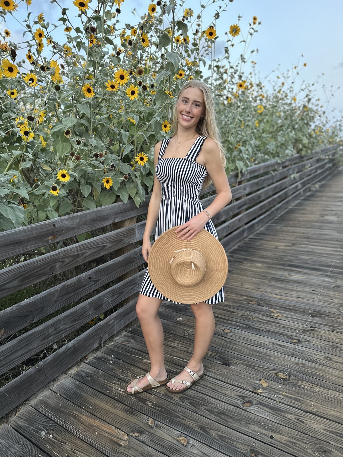 Lacy Dress worn by a woman holding a straw hat, standing on a wooden path surrounded by sunflowers.