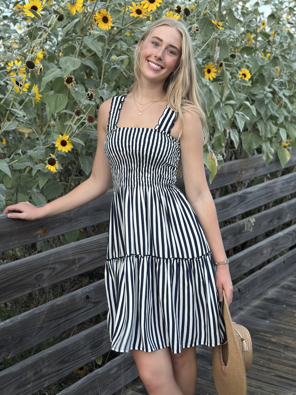 Smiling woman in striped dress by sunflowers, showcasing trendy coastal style from Raggdoll Boutique, Port Aransas.