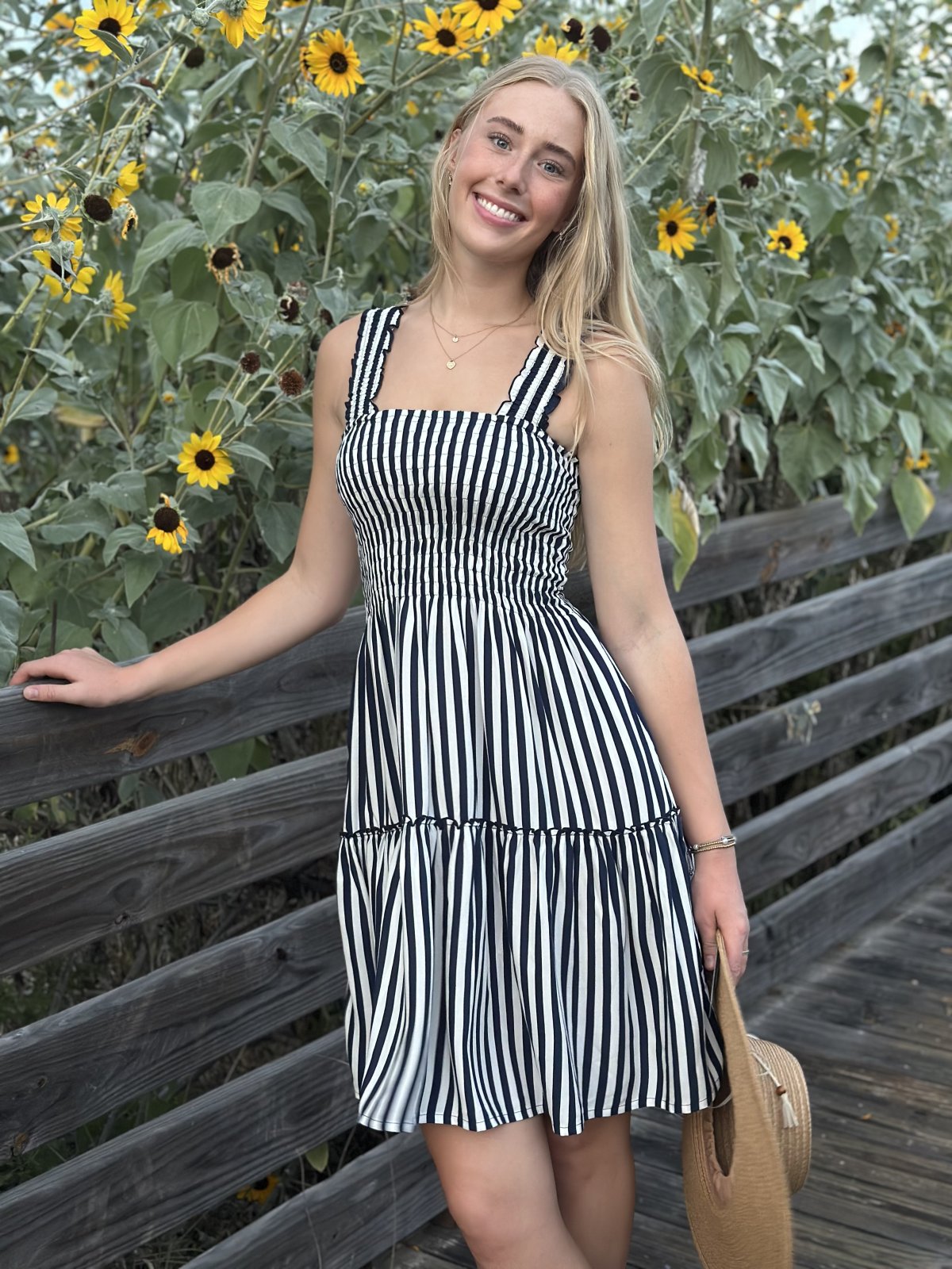 Smiling woman in striped dress by sunflowers, showcasing trendy coastal style from Raggdoll Boutique, Port Aransas.