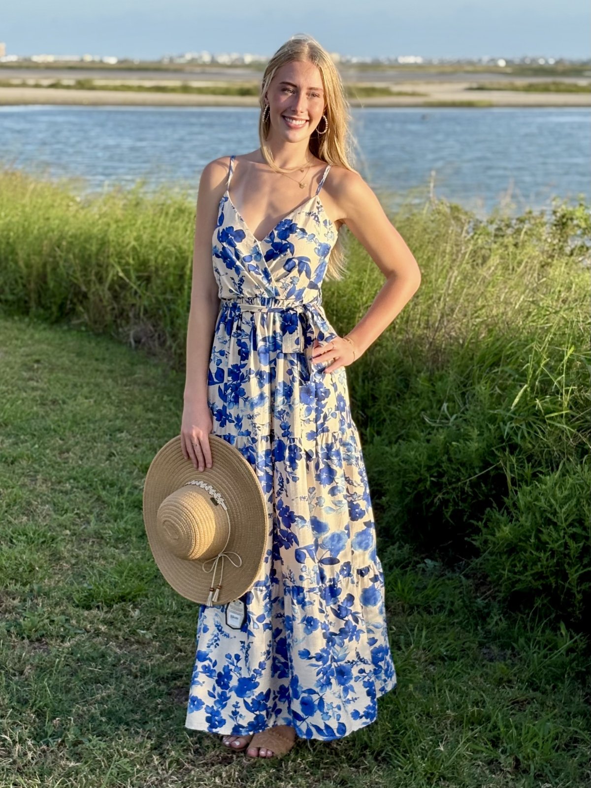 Model wearing a floral print maxi dress by a coastal lake, showcasing curated women's fashion for tropical vacations.