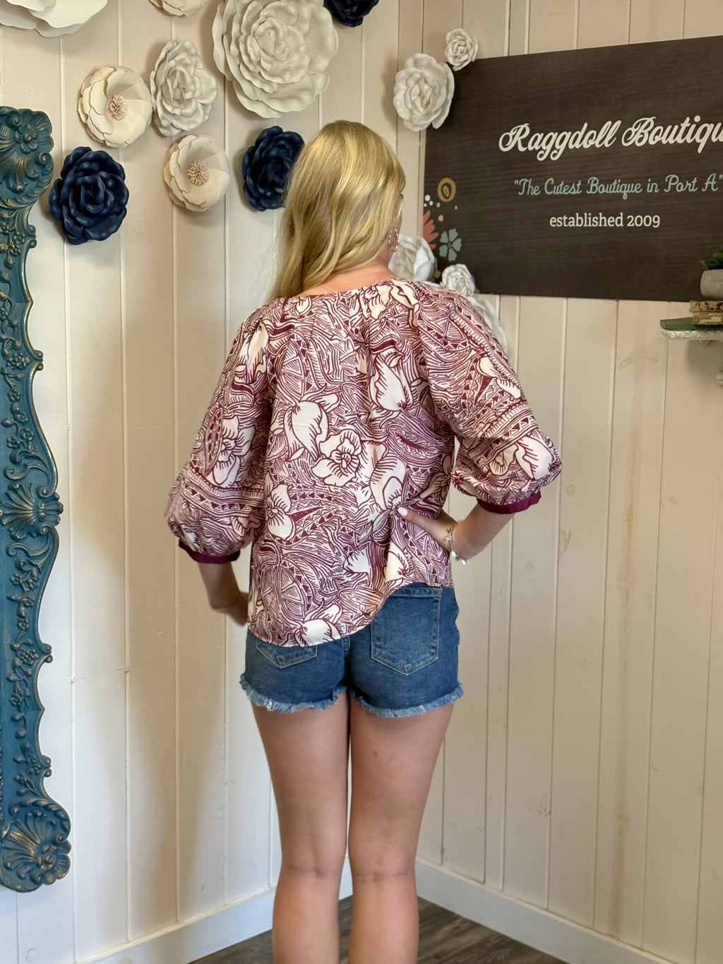 Back view of a woman in a maroon floral Aggie Top with 3/4 sleeves, styled with denim shorts at a boutique.