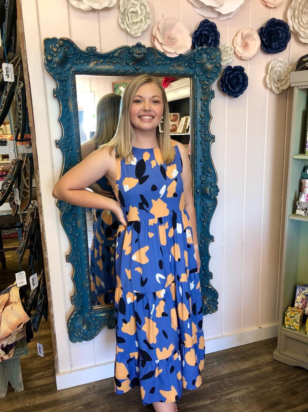 Model wearing a blue print midi dress with back bow in a coastal boutique setting, showcasing curated women's fashion.