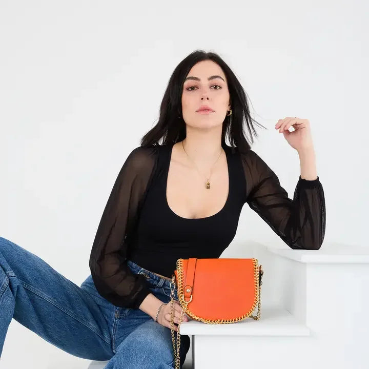 Izzy Crossbody bag in vibrant orange with gold chain trim styled with a sheer black top and high-waisted jeans.